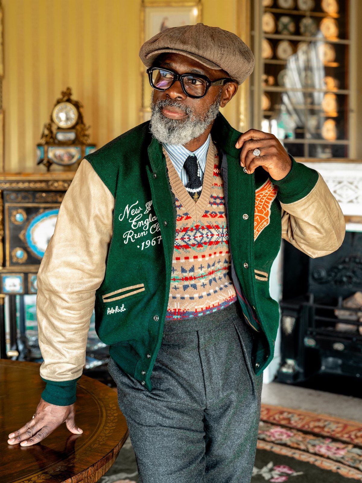 Man in vintage-inspired clothing including a flat cap, patterned sweater vest, and varsity jacket.