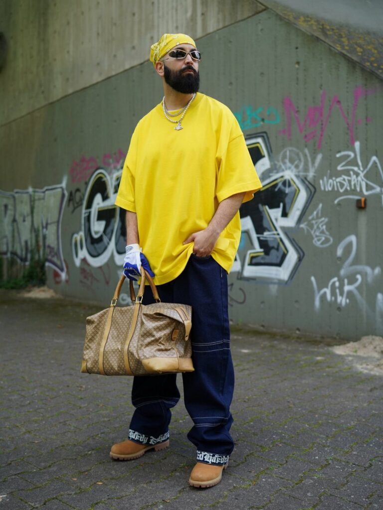 Man in yellow t-shirt and baggy pants wearing tan Timberland boots standing by graffiti.