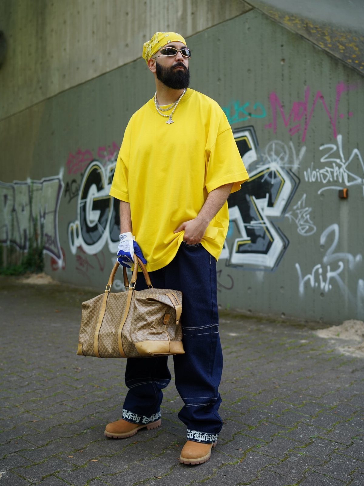Man in yellow t-shirt and baggy pants wearing tan Timberland boots standing by graffiti.