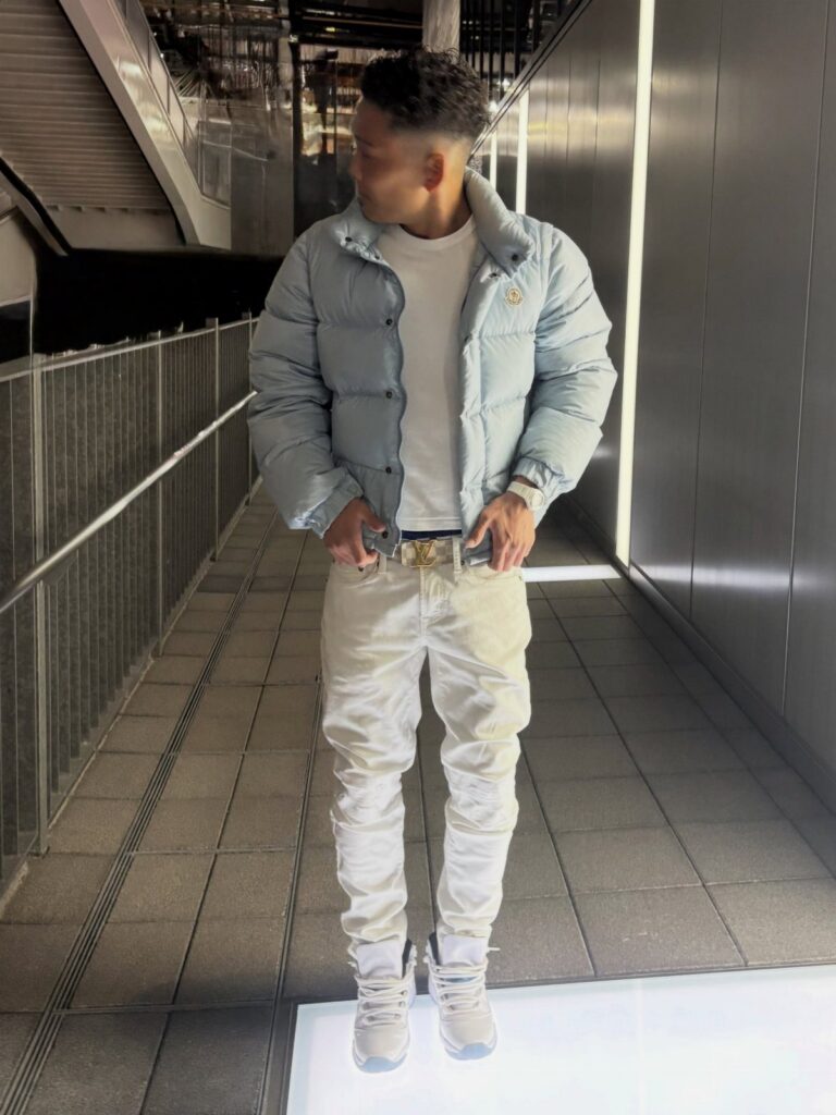 A man posing in white jeans, a light blue puffer jacket, and white sneakers.
