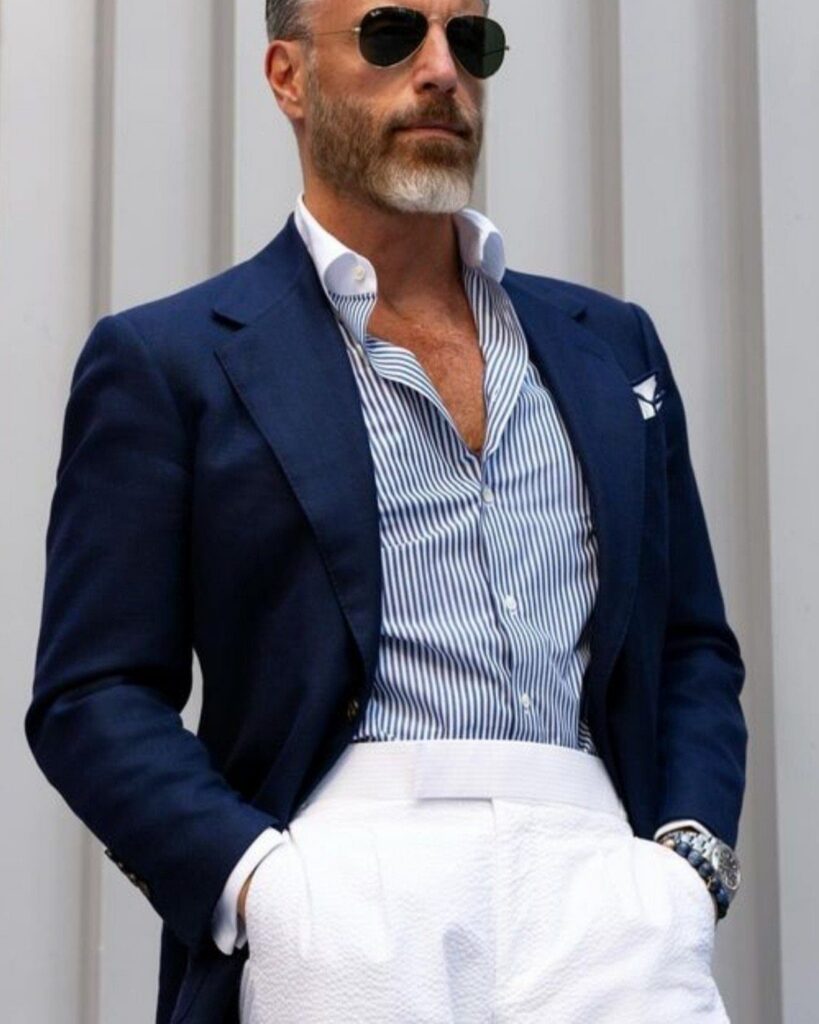 Man in a navy blazer, striped blue and white shirt, and white trousers wearing sunglasses.