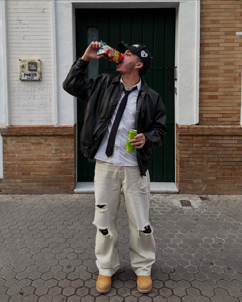 Man in a black leather jacket, white shirt, black tie, and ripped white jeans drinking from a can.