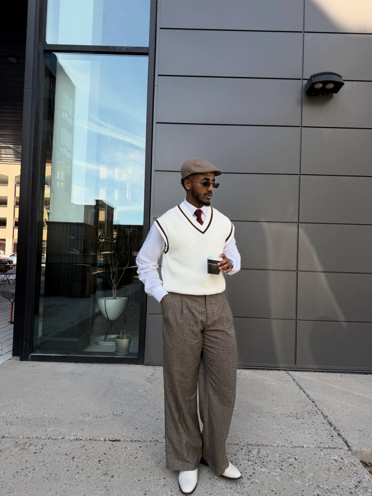 Man wearing a white knitted vest, white shirt, red tie, and patterned wide-leg trousers standing outdoors.