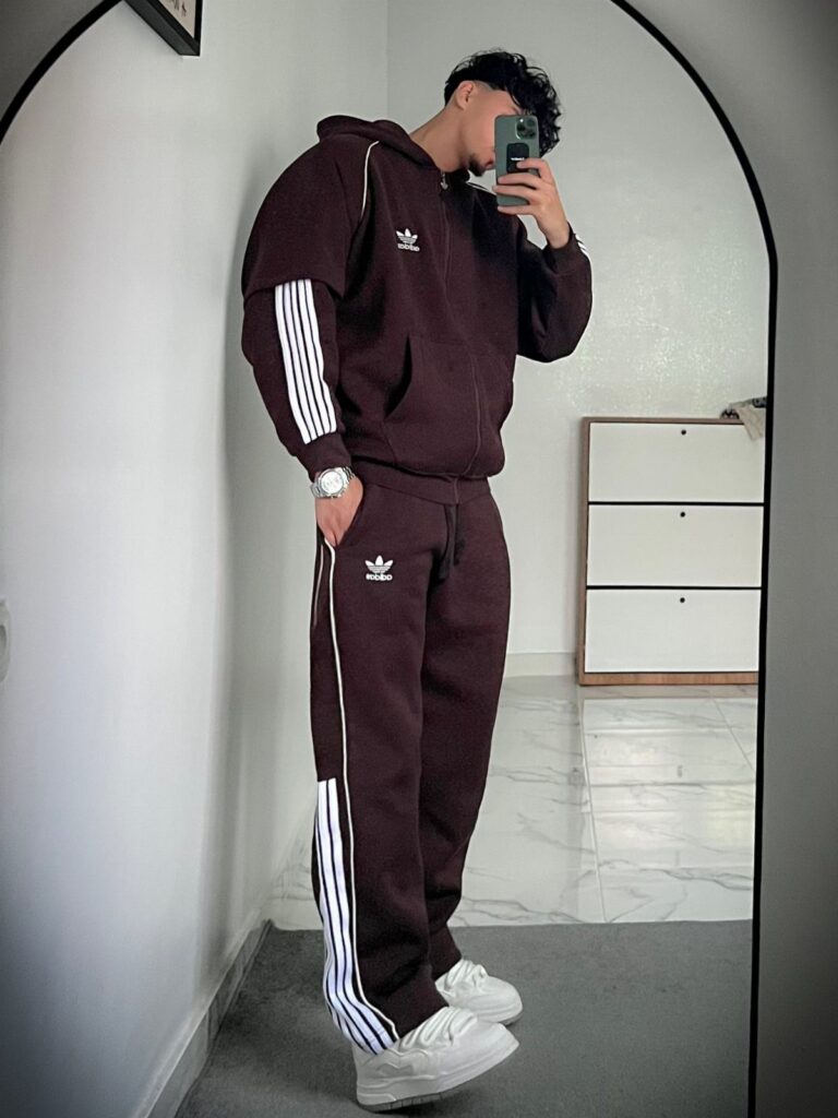 Man in a burgundy Adidas tracksuit and white sneakers posing for a mirror selfie.